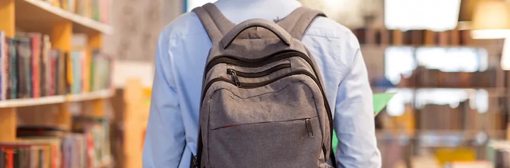 A person in a pale blue shirt with a grey backpack, seen from behind between library bookshelves