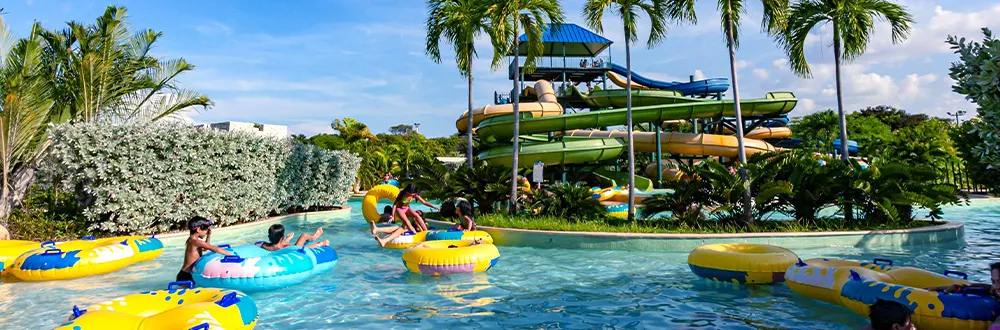 Children and adults floating on yellow and blue rubber rings along a lazy river lined with palm trees, with water slides behind