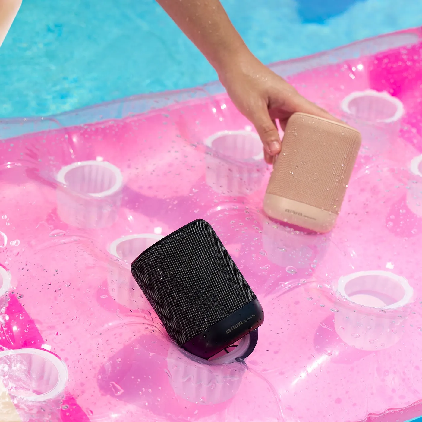 A hand reaching for a pink AIWA speaker beside a black AIWA speaker on a wet pink pool float