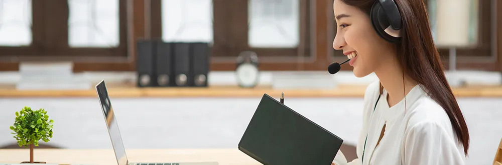 A smiling young woman in a headset with a microphone holding a dark green notebook beside an open laptop at a wooden desk