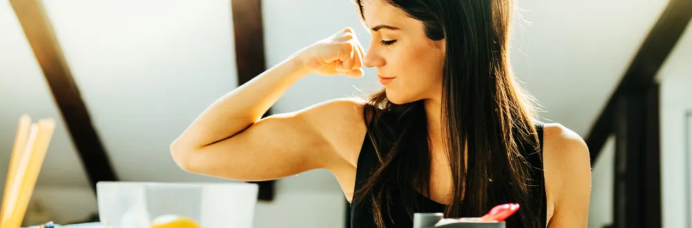 A woman with long dark hair in a black sleeveless top flexing her arm in a sunlit room with wooden roof beams