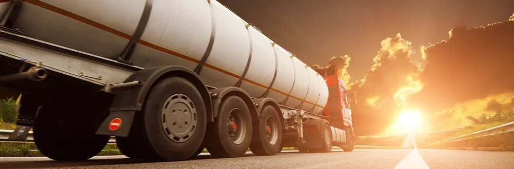 A silver tanker truck with a red cab driving along a highway towards a low sun, seen from behind its rear wheels