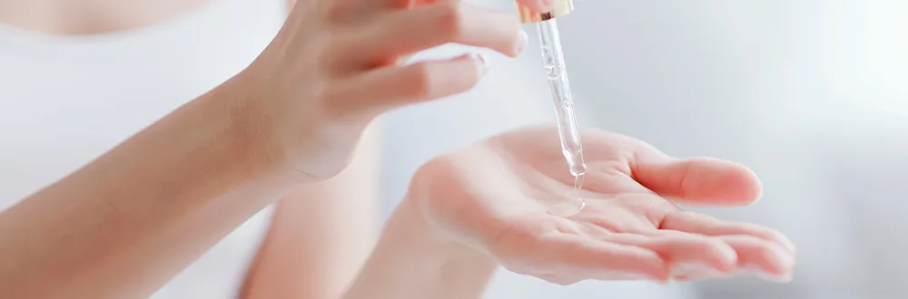 A hand squeezing a glass dropper of clear serum onto the open palm of the other hand