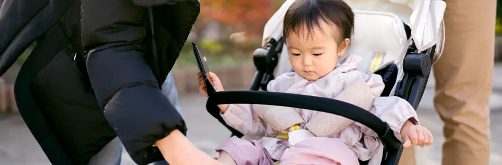 A toddler in a pushchair holding a phone, an adult in a black jacket beside it