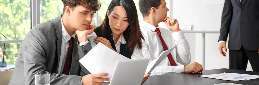 Three young people in office clothes reading papers and a laptop at a meeting table, a man in a dark suit standing beside them