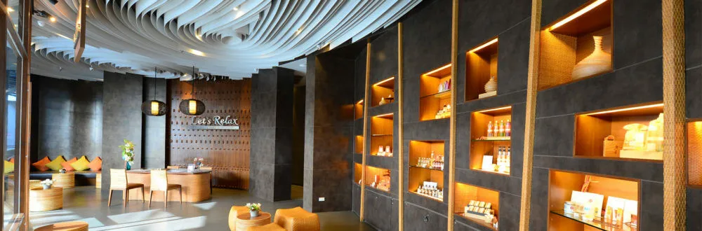 A spa reception with a Let's Relax sign on a wooden wall, cushioned benches, rattan seats and lit shelves of spa products
