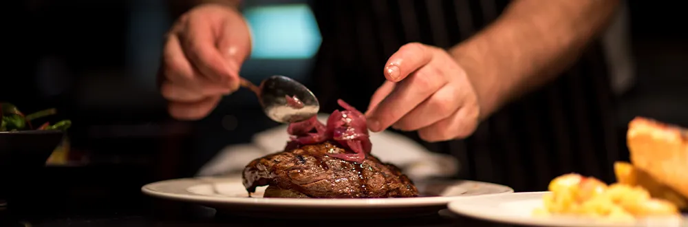 A cook spooning red onion onto a grilled steak on a white plate