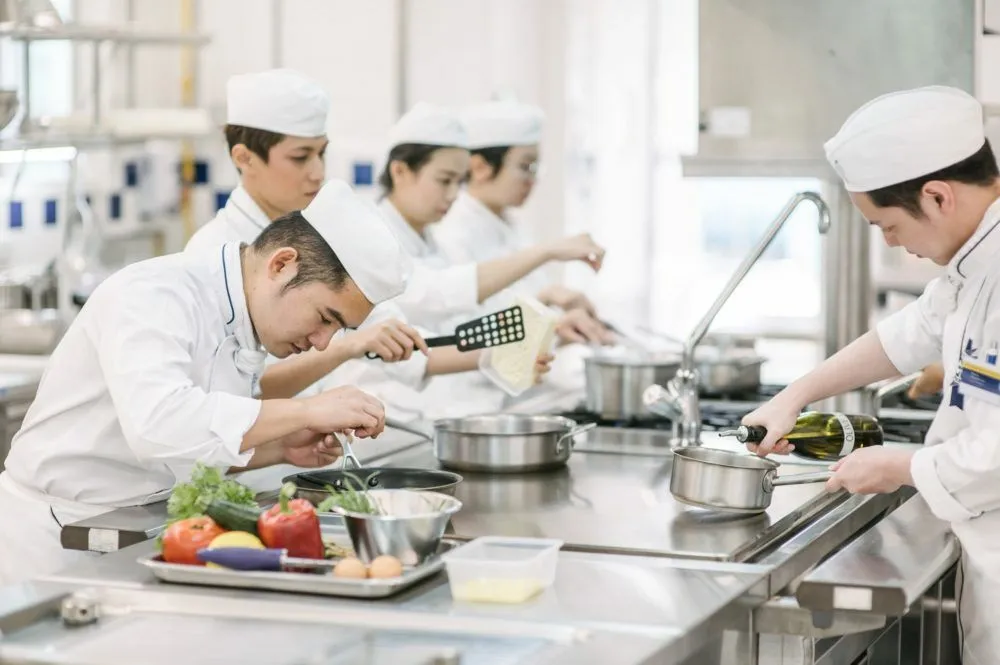 Cooks in white jackets and caps preparing food at a steel counter, with a tray of vegetables, pans and saucepans in front of them