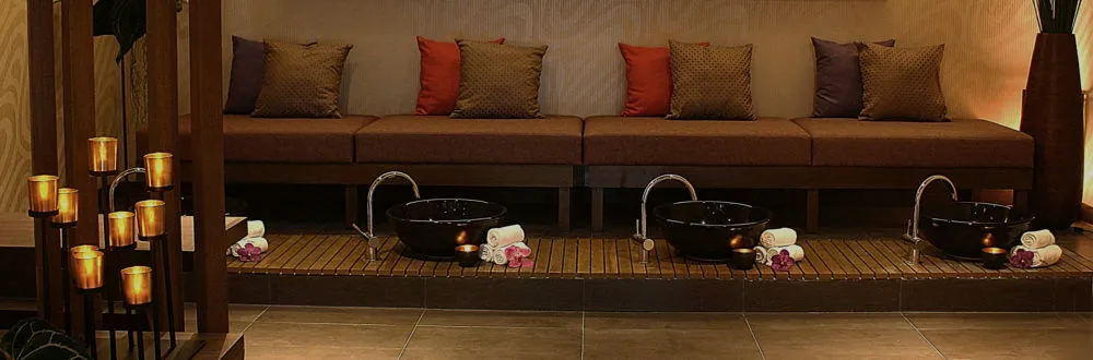 A row of black foot basins with folded towels on a wooden platform in front of a long sofa with cushions, beside lit candles