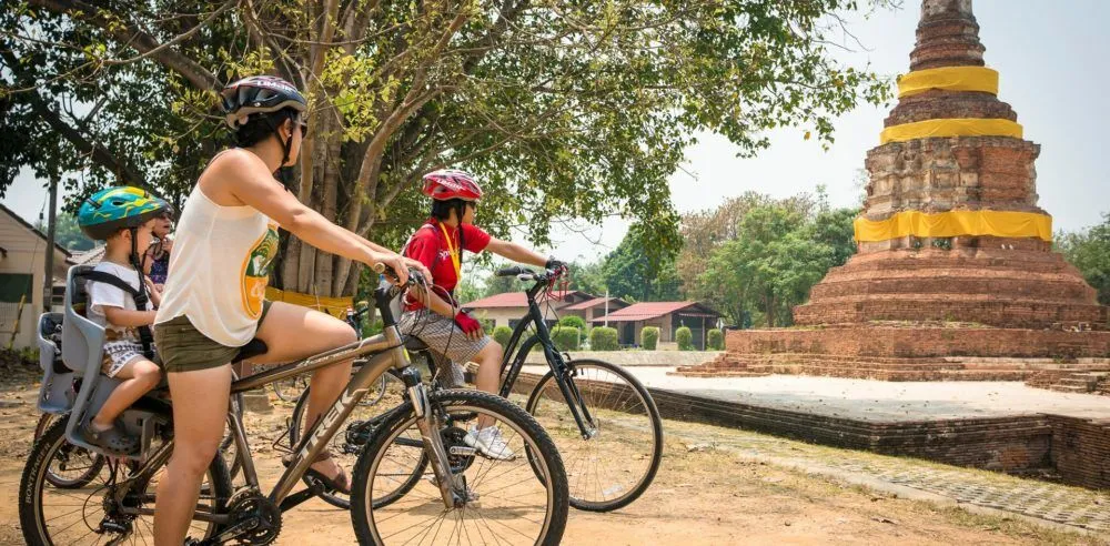 A woman and 2 children on bicycles beside an old brick stupa wrapped with yellow cloth